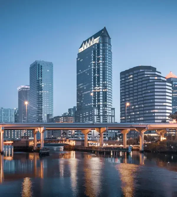 Downtown skyline at dusk, featuring modern skyscrapers and a bridge reflected in the calm water below.