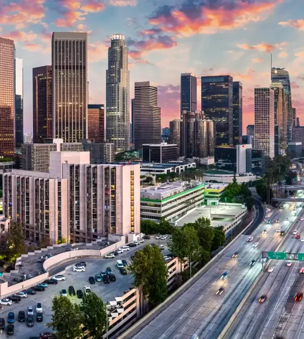 Sunset over downtown Los Angeles, featuring modern skyscrapers, lush greenery, and a busy highway lined with vehicles.