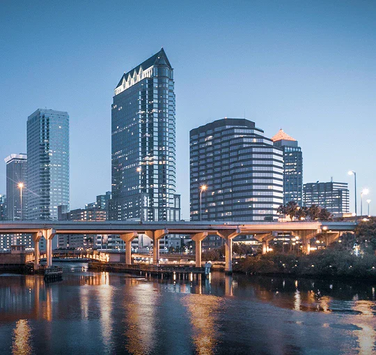 Downtown skyline at dusk, featuring modern skyscrapers and a bridge reflected in the calm water below.