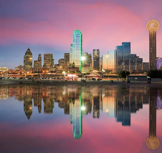 Dallas skyline shimmering at dusk, reflecting on calm water under a vibrant pink and purple sky, highlighted by illuminated buildings.
